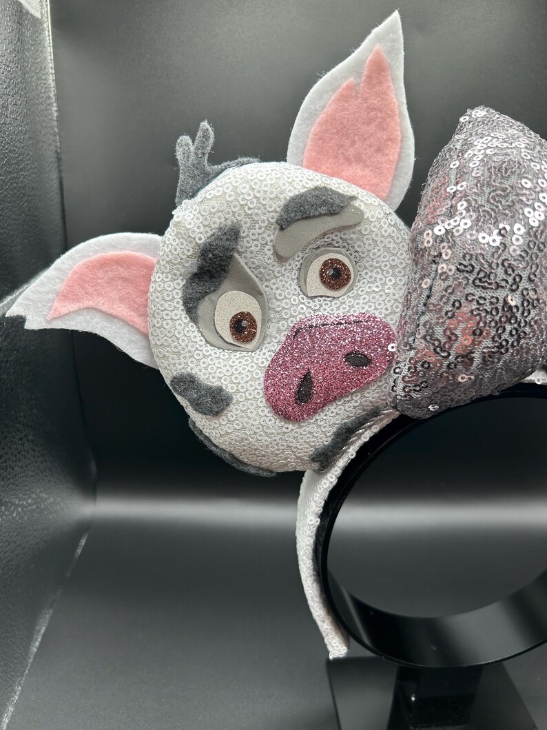 May include: A headband featuring a character face covered in white sequins with grey felt patches, pink felt inner ears, and a glittery pink snout. The character has brown eyes and grey felt eyebrows. A silver sequined bow sits to the right side.