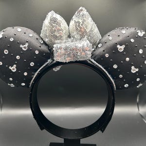 May include: Black satin mouse ear headband with silver sequin bow and sparkling crystal accents. The ears are adorned with scattered crystals and Mickey Mouse head-shaped gems. A fun accessory for themed events.