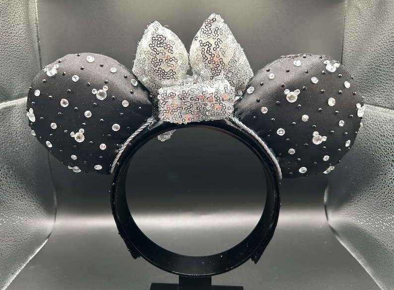 May include: Black satin mouse ear headband with silver sequin bow and sparkling crystal accents. The ears are adorned with scattered crystals and Mickey Mouse head-shaped gems. A fun accessory for themed events.