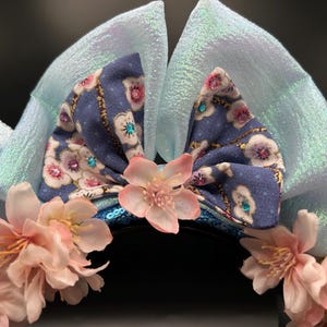 Peut inclure: Serre-t&ecirc;te avec deux grandes oreilles en tissu bleu clair iris&eacute; et un n&oelig;ud central en tissu bleu &agrave; motif floral blanc et rose. Le n&oelig;ud est orn&eacute; de petits strass bleus et roses. Des fleurs artificielles rose p&acirc;le sont fix&eacute;es sur le bandeau.