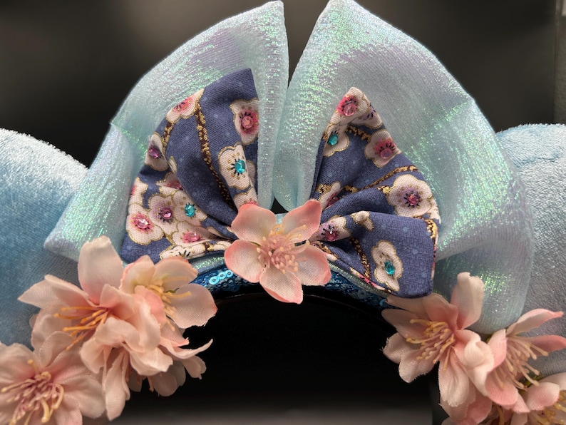 Peut inclure: Serre-t&ecirc;te avec deux grandes oreilles en tissu bleu clair iris&eacute; et un n&oelig;ud central en tissu bleu &agrave; motif floral blanc et rose. Le n&oelig;ud est orn&eacute; de petits strass bleus et roses. Des fleurs artificielles rose p&acirc;le sont fix&eacute;es sur le bandeau.