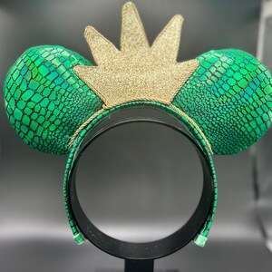 Op de afbeelding: Een hoofdband met muisoren met groene oren met slangenleerpatroon en een gouden glitterkroon. De zwarte hoofdband heeft een groen slangenleerpatroon. De oren zijn rond en gewatteerd.