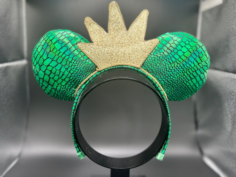Op de afbeelding: Een hoofdband met muisoren met groene oren met slangenleerpatroon en een gouden glitterkroon. De zwarte hoofdband heeft een groen slangenleerpatroon. De oren zijn rond en gewatteerd.