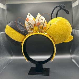 May include: A pair of yellow and black bee-themed Minnie Mouse ears with a black headband and a bow with a honeycomb pattern.