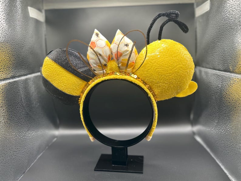 May include: A pair of yellow and black bee-themed Minnie Mouse ears with a black headband and a bow with a honeycomb pattern.