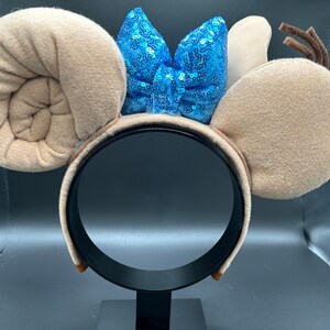 May include: Tan mouse ears headband with a blue sequined bow. The ears are shaped like a spiral and a rounded ear.