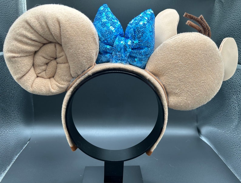 May include: Tan mouse ears headband with a blue sequined bow. The ears are shaped like a spiral and a rounded ear.