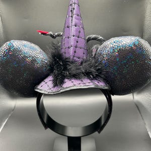 May include: A witch-themed headband with black ears and a purple witch hat. The hat is adorned with sequins and a black feather. The ears have a textured, iridescent finish. The headband is black and sits on a black display stand.