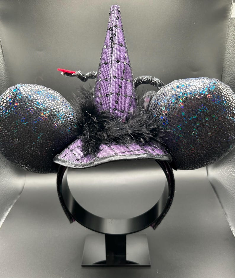May include: A witch-themed headband with black ears and a purple witch hat. The hat is adorned with sequins and a black feather. The ears have a textured, iridescent finish. The headband is black and sits on a black display stand.