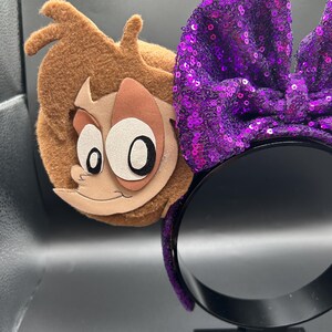 Op de afbeelding: Een haarband met een grote paarse lovertjesstrik en een bruin vilten oor met een applicatie van een cartoongezicht. Het personage heeft een lichtbruine huid, grote witte ogen met zwarte pupillen en bruin haar. De haarband is bedekt met paarse lovertjes.