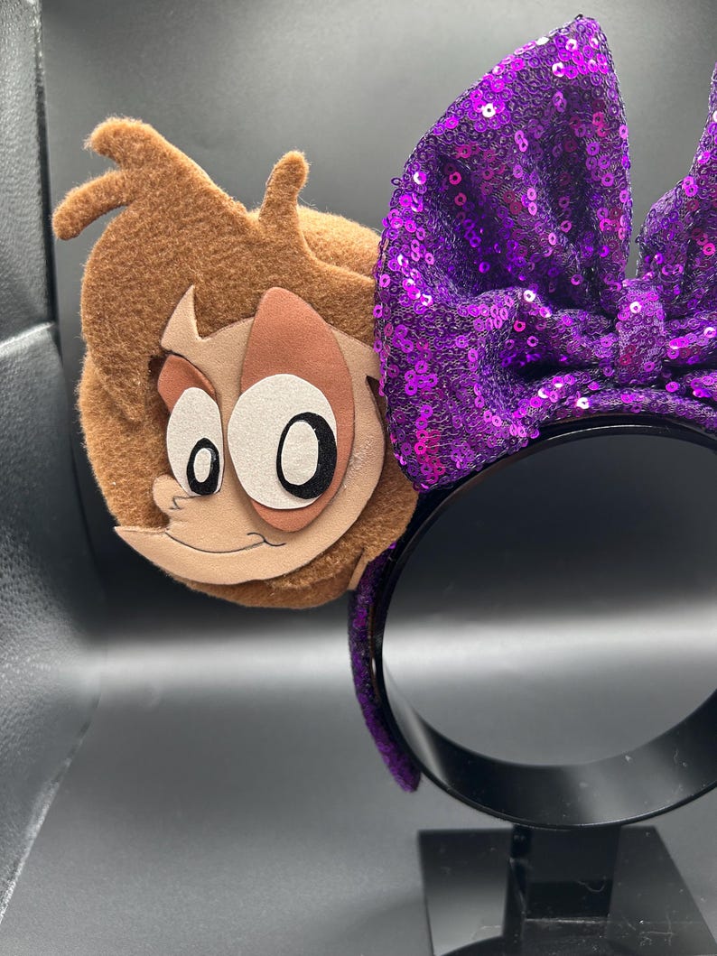Op de afbeelding: Een haarband met een grote paarse lovertjesstrik en een bruin vilten oor met een applicatie van een cartoongezicht. Het personage heeft een lichtbruine huid, grote witte ogen met zwarte pupillen en bruin haar. De haarband is bedekt met paarse lovertjes.