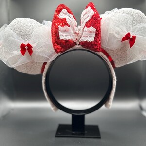 May include: White mouse ears with a red and white bow with lace and sequins. The ears are on a black stand.