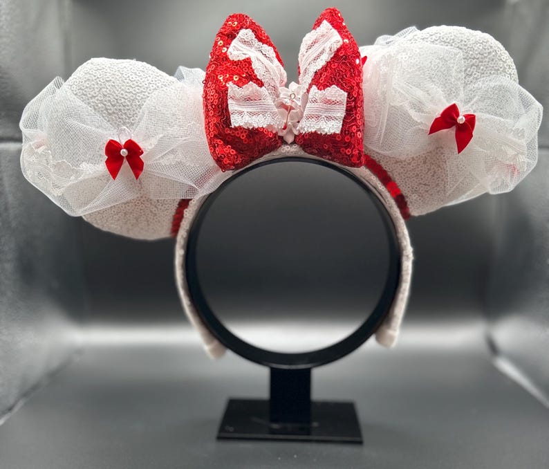 May include: White mouse ears with a red and white bow with lace and sequins. The ears are on a black stand.