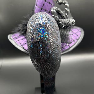 May include: A decorative witch hat headband with a purple and black color scheme. The hat features a textured, iridescent design on the front, a purple brim, and a black feather accent. The hat is mounted on a black headband.