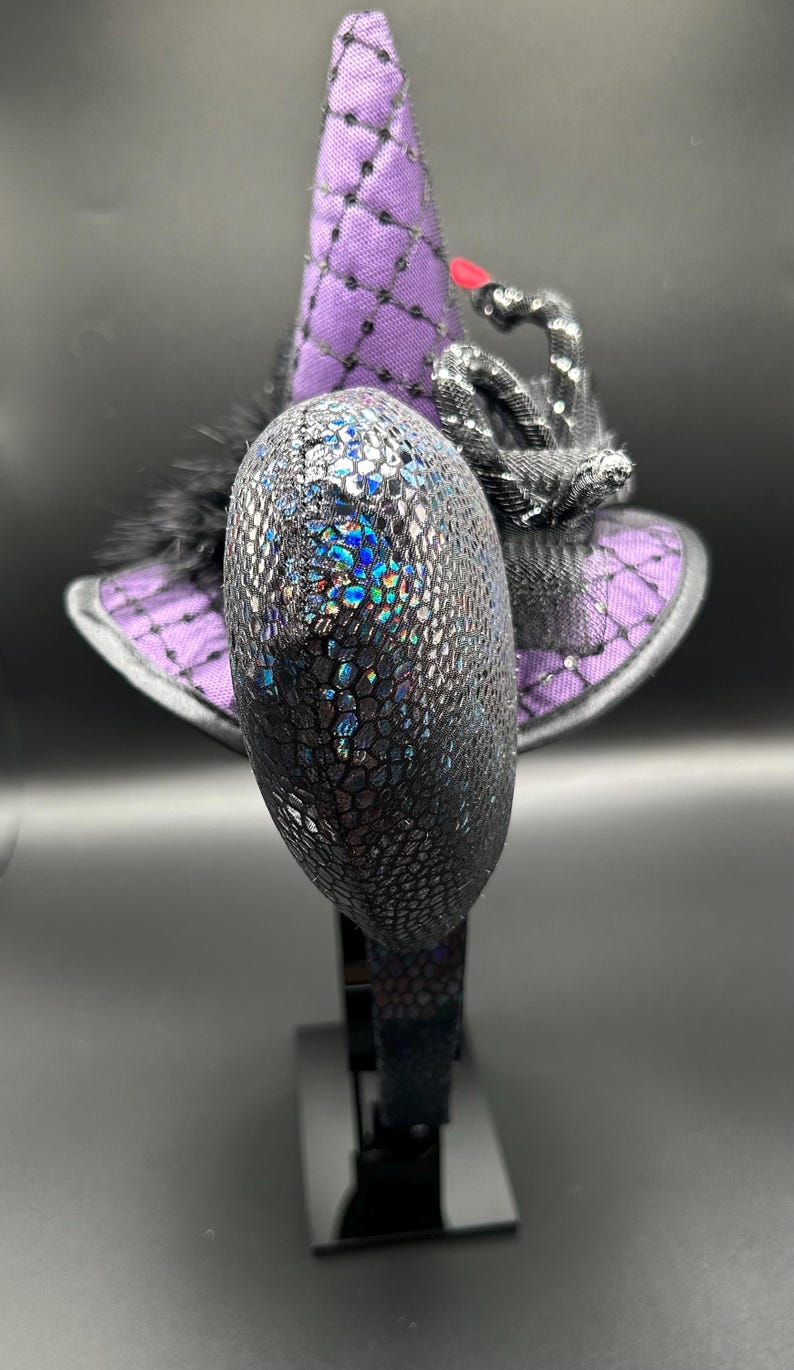 May include: A decorative witch hat headband with a purple and black color scheme. The hat features a textured, iridescent design on the front, a purple brim, and a black feather accent. The hat is mounted on a black headband.