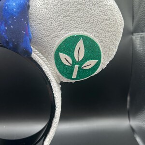 May include: White sequined mouse ears with a green and white leaf emblem. A blue bow with a galaxy print is attached to the headband. The headband is black.