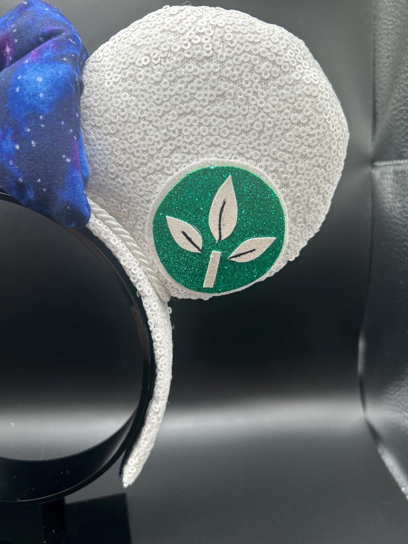 May include: White sequined mouse ears with a green and white leaf emblem. A blue bow with a galaxy print is attached to the headband. The headband is black.