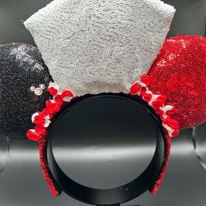 May include: A headband featuring two circular ears covered in black and red sequins, with a central white sequined bow. Small red and white fabric roses are arranged along the base of the ears and bow, and a small crystal accent is on the black ear.