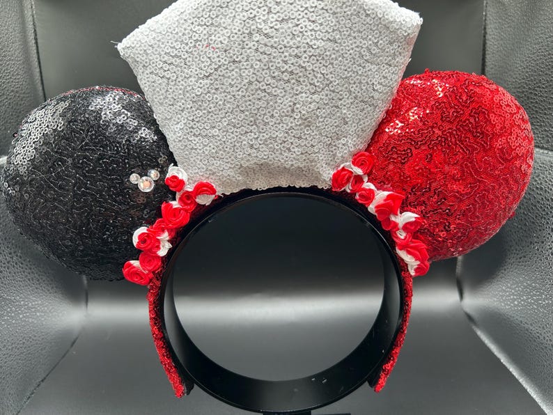 May include: A headband featuring two circular ears covered in black and red sequins, with a central white sequined bow. Small red and white fabric roses are arranged along the base of the ears and bow, and a small crystal accent is on the black ear.