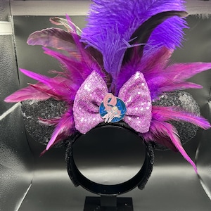 May include: A headband featuring two black sequined ears, a central light purple sequined bow, and a circular pin depicting a pink character. The design is accented with vibrant purple and dark magenta feathers extending outward from the center.