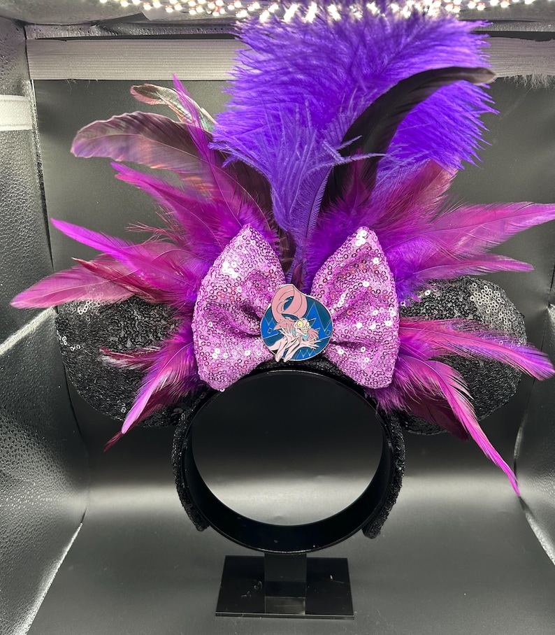 May include: A headband featuring two black sequined ears, a central light purple sequined bow, and a circular pin depicting a pink character. The design is accented with vibrant purple and dark magenta feathers extending outward from the center.