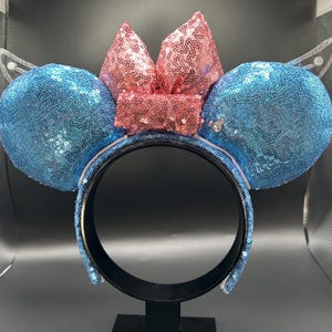 May include: A sequined headband with large blue ears and a pink bow. The ears and bow are covered in shimmering sequins. The headband has a black base and is decorated with blue sequins.