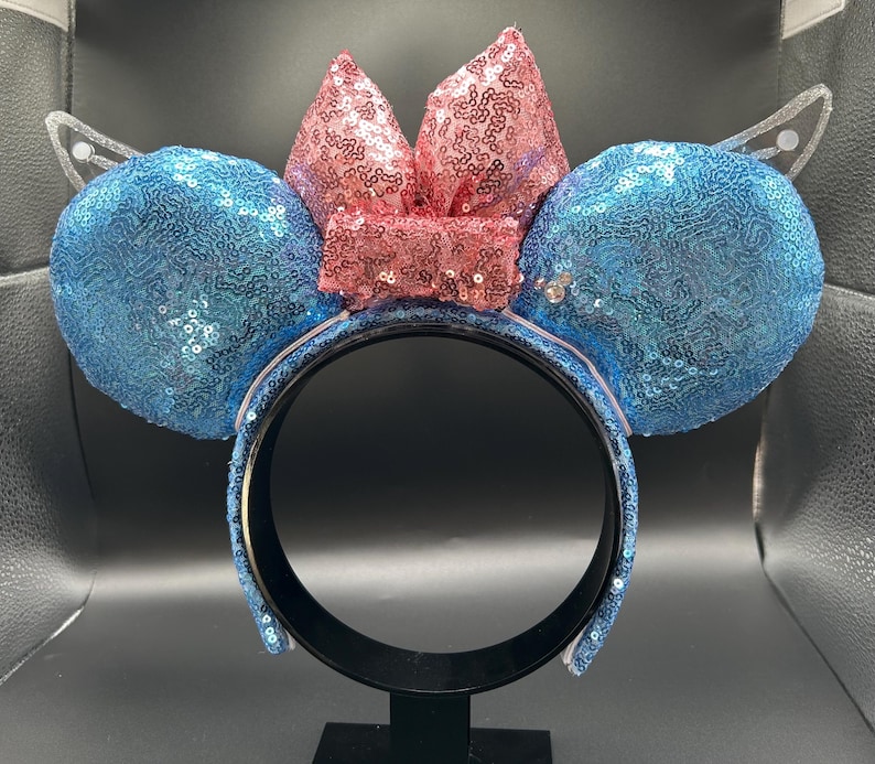 May include: A sequined headband with large blue ears and a pink bow. The ears and bow are covered in shimmering sequins. The headband has a black base and is decorated with blue sequins.