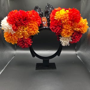 May include: A black headband featuring two large circular clusters of artificial flowers in shades of red, orange, and white. Between the floral clusters sits a black sequined bow with a central orange sequined accent piece.