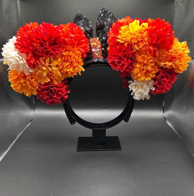 May include: A black headband featuring two large circular clusters of artificial flowers in shades of red, orange, and white. Between the floral clusters sits a black sequined bow with a central orange sequined accent piece.