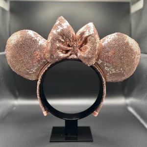 May include: Rose gold sequined Minnie Mouse ears headband with a matching bow. The ears and bow are covered in small, shiny sequins. The headband is black and has a simple design.