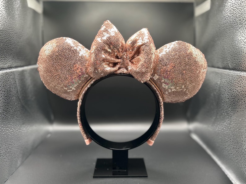 May include: Rose gold sequined Minnie Mouse ears headband with a matching bow. The ears and bow are covered in small, shiny sequins. The headband is black and has a simple design.