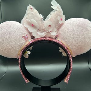 May include: A pale pink headband with two fluffy ears. The headband features a sequined band, white flowers, and a sheer bow. The ears are decorated with small, pink sequins. A decorative accessory.