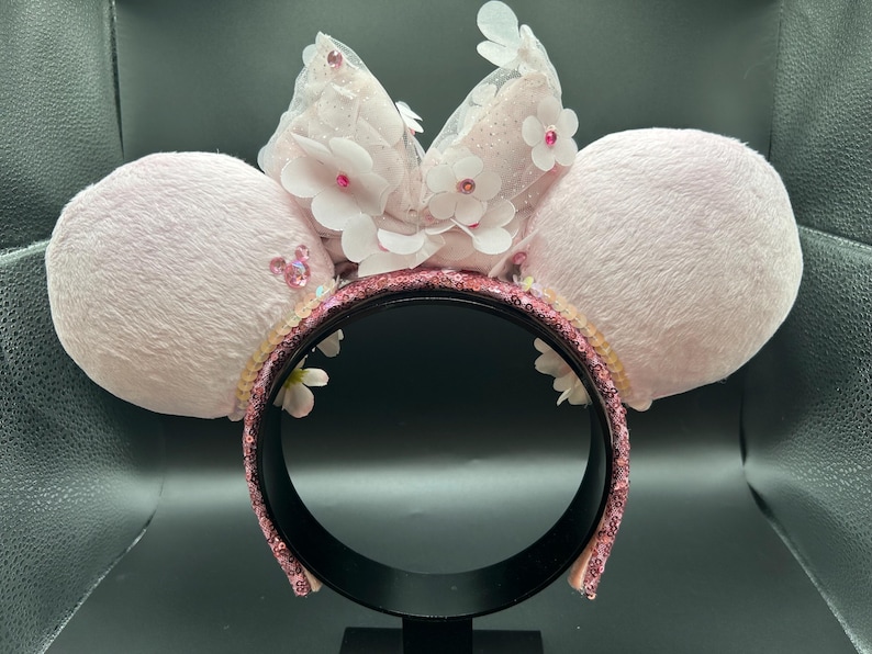 May include: A pale pink headband with two fluffy ears. The headband features a sequined band, white flowers, and a sheer bow. The ears are decorated with small, pink sequins. A decorative accessory.