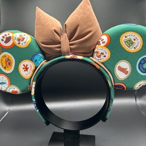 May include: Green mouse ears headband with a brown bow. The ears are covered in a green fabric with colourful patches that say "Campfire Chef", "Take Life", "Crafty Kid", "Shine Bright", and more.