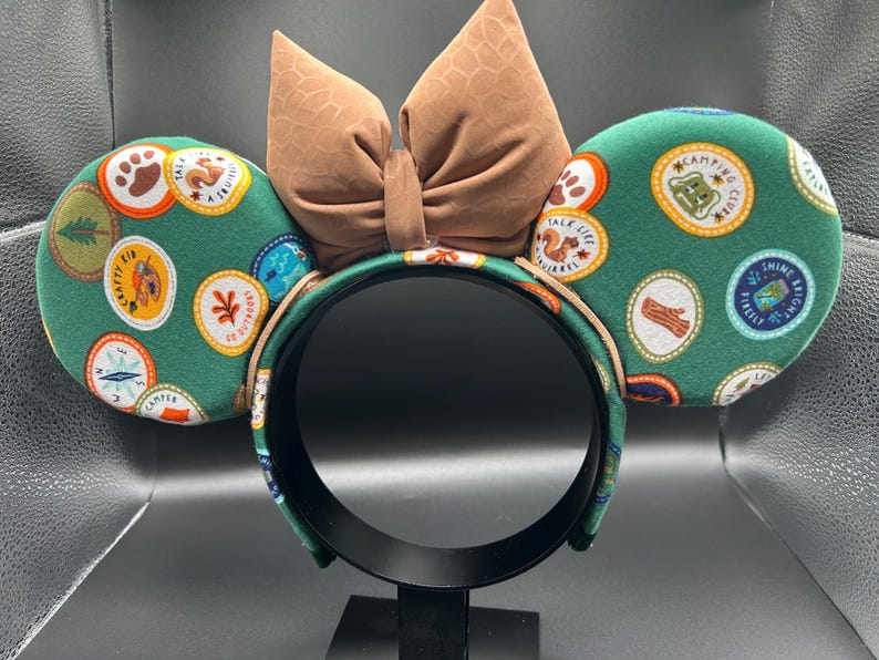 May include: Green mouse ears headband with a brown bow. The ears are covered in a green fabric with colourful patches that say "Campfire Chef", "Take Life", "Crafty Kid", "Shine Bright", and more.