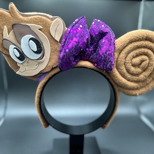 Può includere: Fascia per capelli con orecchie di scimmia marroni e un fiocco di paillettes viola. Il viso della scimmia è marrone con occhi bianchi e un naso marrone. La fascia è nera.