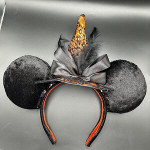 Puede incluir: Una diadema con dos grandes orejas circulares de terciopelo negro y un sombrero de bruja puntiagudo en el centro. El sombrero est&aacute; hecho de tela naranja con una capa de encaje negro, acentuado por un lazo de sat&eacute;n negro y plumas negras.
