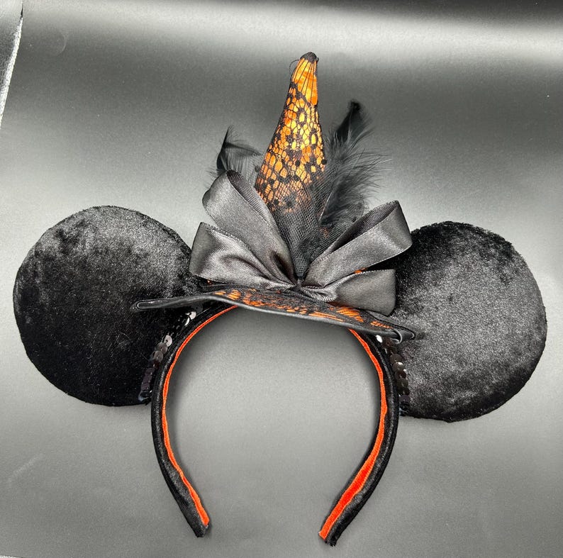 Puede incluir: Una diadema con dos grandes orejas circulares de terciopelo negro y un sombrero de bruja puntiagudo en el centro. El sombrero est&aacute; hecho de tela naranja con una capa de encaje negro, acentuado por un lazo de sat&eacute;n negro y plumas negras.