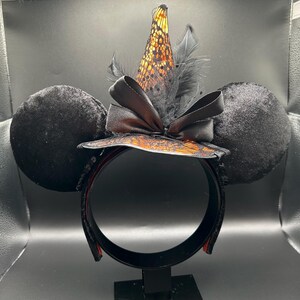 Puede incluir: Una diadema con dos grandes orejas redondas de terciopelo negro y un sombrero de bruja central. El sombrero es naranja con una capa de encaje negro, acentuado por un lazo de sat&eacute;n negro y plumas negras. La diadema es negra con un forro interior naranja.