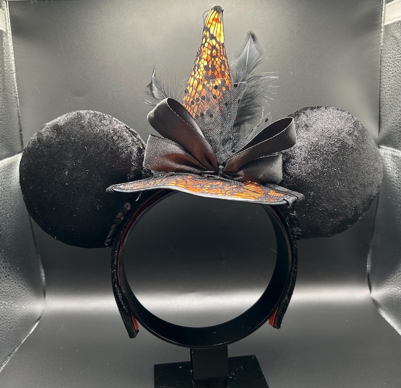 Puede incluir: Una diadema con dos grandes orejas redondas de terciopelo negro y un sombrero de bruja central. El sombrero es naranja con una capa de encaje negro, acentuado por un lazo de sat&eacute;n negro y plumas negras. La diadema es negra con un forro interior naranja.