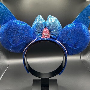 May include: A blue sequined headband with large, round ears and wing-shaped accents. A light blue bow with a pink center and a small jewel is centered between the ears. The headband is black with a white trim.