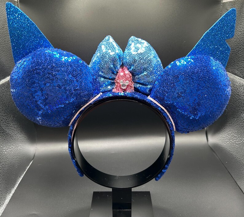 May include: A blue sequined headband with large, round ears and wing-shaped accents. A light blue bow with a pink center and a small jewel is centered between the ears. The headband is black with a white trim.