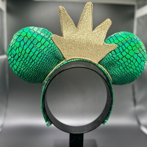 Op de afbeelding: Een groen en zwart patroon hoofdband met twee ronde oren en een gouden glitter kroon. De oren hebben een reptielachtige textuur. De kroon is gemaakt van gouden glitter en is met gouden touw aan de hoofdband bevestigd.