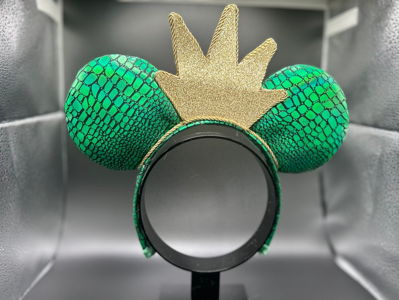 Op de afbeelding: Een groen en zwart patroon hoofdband met twee ronde oren en een gouden glitter kroon. De oren hebben een reptielachtige textuur. De kroon is gemaakt van gouden glitter en is met gouden touw aan de hoofdband bevestigd.