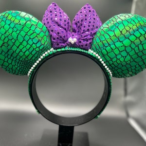 Op de afbeelding: Een zwarte hoofdband met groene, geschubde oren en een paarse paillettenstrik. De oren zijn rond en de strik is gecentreerd. Een rij witte parels omzoomt de hoofdband. Het ontwerp doet denken aan een zeemeermin of zeewezen.