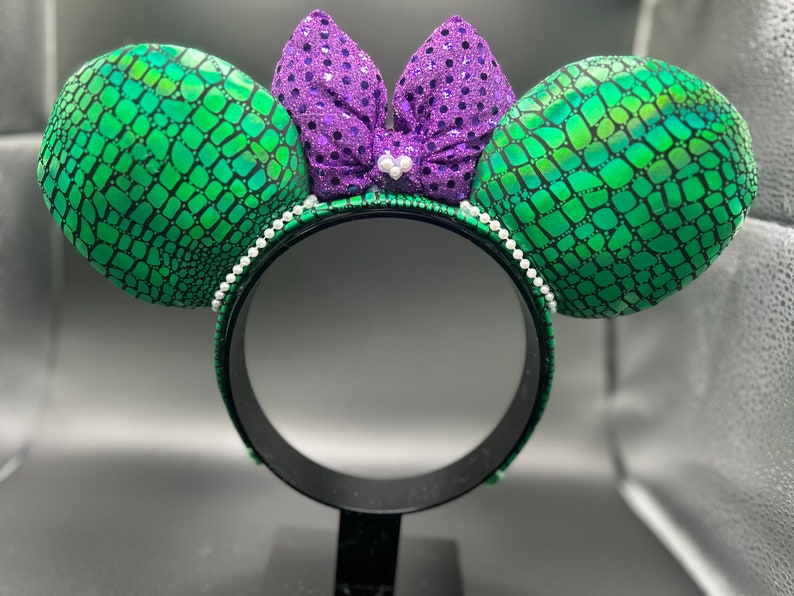 Op de afbeelding: Een zwarte hoofdband met groene, geschubde oren en een paarse paillettenstrik. De oren zijn rond en de strik is gecentreerd. Een rij witte parels omzoomt de hoofdband. Het ontwerp doet denken aan een zeemeermin of zeewezen.