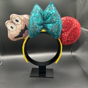 May include: A whimsical headband featuring a teal sequined bow, a red sequined ear, and a brown character with large eyes. The headband has a yellow band and is displayed on a black stand.