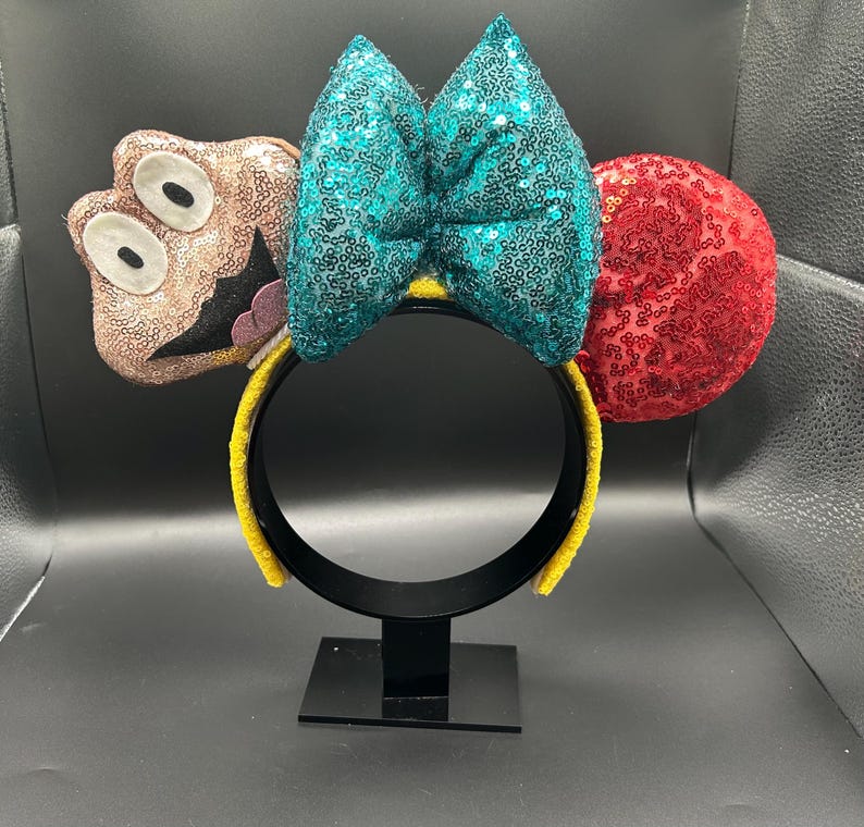 May include: A whimsical headband featuring a teal sequined bow, a red sequined ear, and a brown character with large eyes. The headband has a yellow band and is displayed on a black stand.