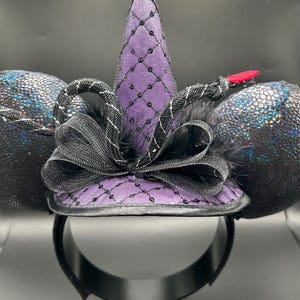 May include: A black headband with attached ears and a witch hat design. The ears are a textured, iridescent black. The purple witch hat is adorned with a black net pattern and a black bow. A red accent is on the side.