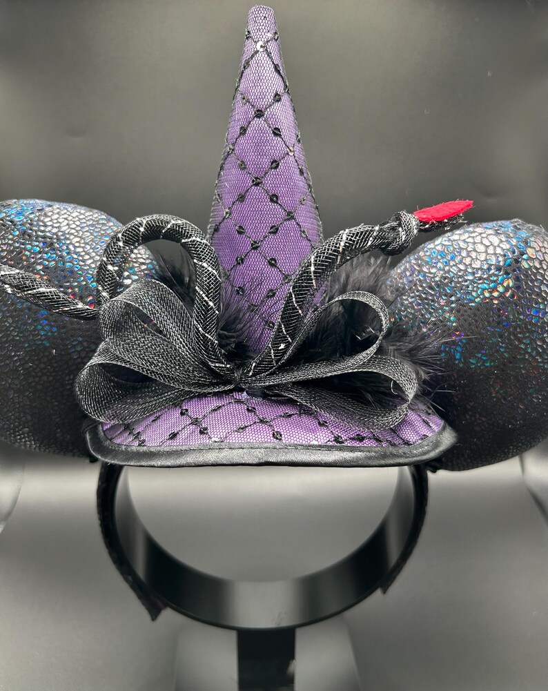 May include: A black headband with attached ears and a witch hat design. The ears are a textured, iridescent black. The purple witch hat is adorned with a black net pattern and a black bow. A red accent is on the side.